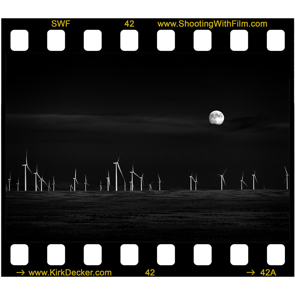 Photograph by Kirk Decker.  A full Moon rises behind field of wind turbines.  The sky is very dark with a few clouds.  The wind mills are illuminated by a setting sun and are stark white against the dark sky giving the impression of alien flowers harvesting Moon light.
