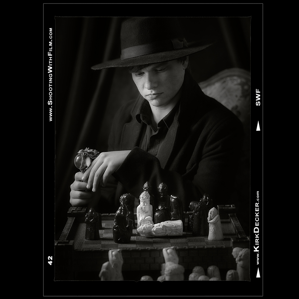 Portrait by Kirk Decker of a young man dressed in black with a black hat and dragon cane looking down at a toppled queen on a chessboard.  The photo is dark and moody the chess pieces resemble the medieval characters found in the Lewis chess set.