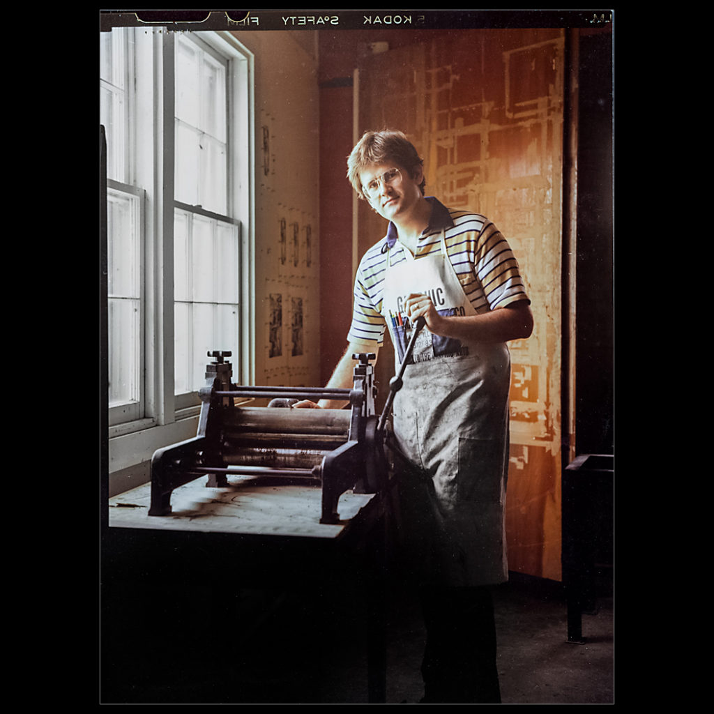 Large format portrait of artist Brent Pulsipher taken by Kansas City photographer Kirk Decker.  Photograph taken in 1980. Brent is window lit and standing by a etching press.